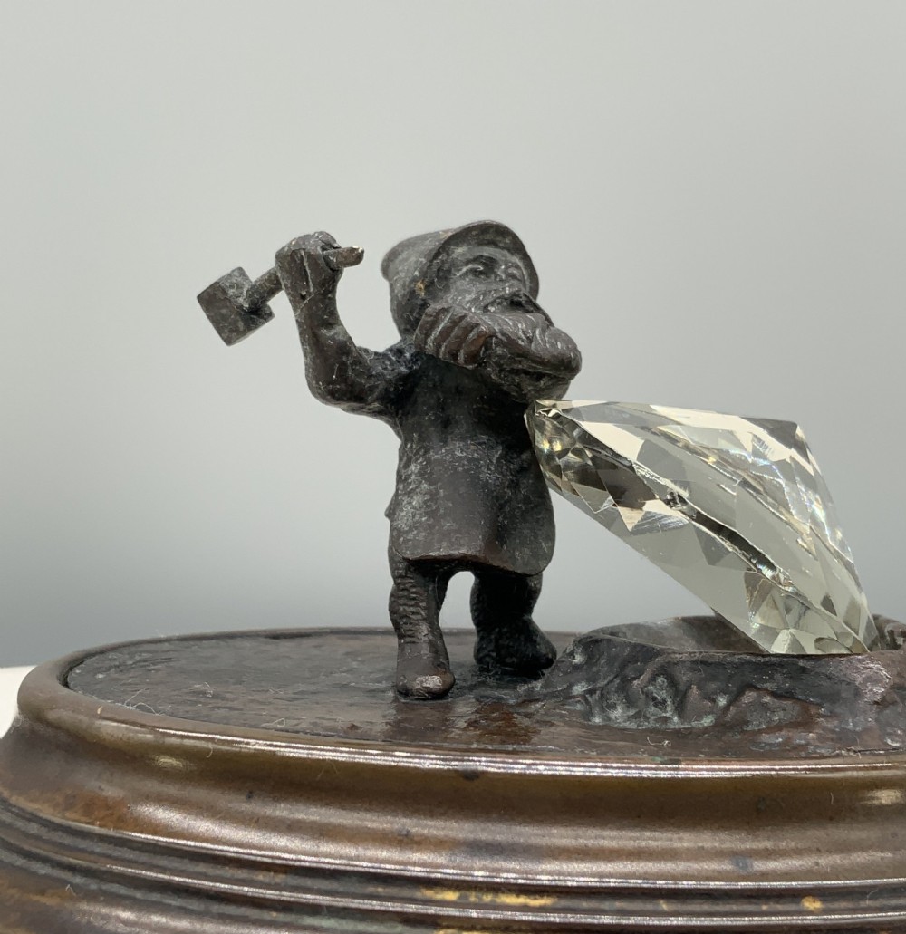 A small dark bronze figurine of a bearded dwarf miner holding a hammer overhead, standing on a molded base next to a faceted cut crystal gem. View III