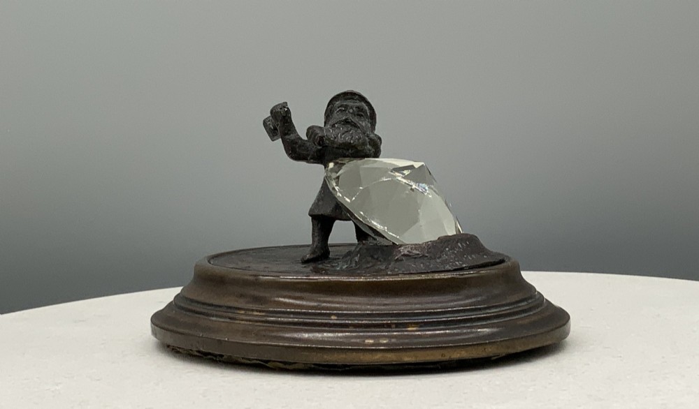 A small dark bronze figurine of a bearded dwarf miner holding a hammer overhead, standing on a molded base next to a faceted cut crystal gem. View IV