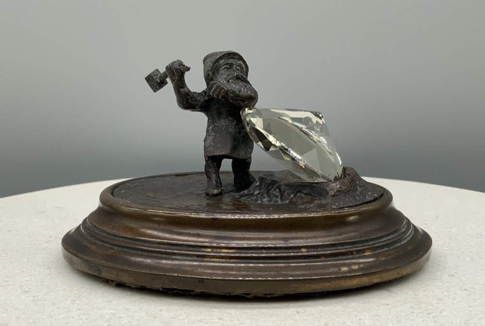 A small dark bronze figurine of a bearded dwarf miner holding a hammer overhead, standing on a molded base next to a faceted cut crystal gem. View V