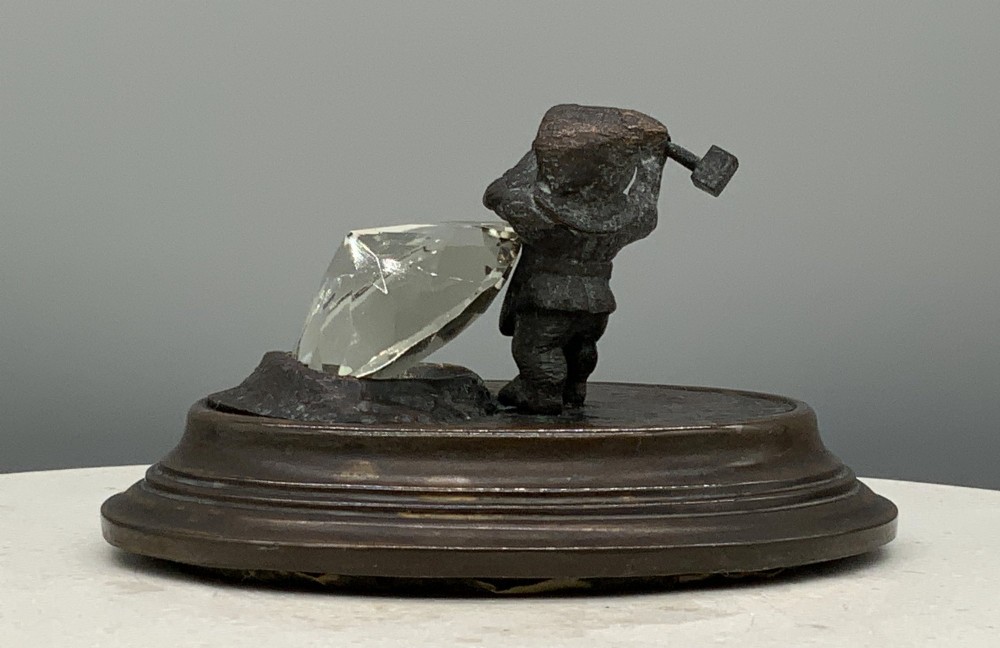 A small dark bronze figurine of a bearded dwarf miner holding a hammer overhead, standing on a molded base next to a faceted cut crystal gem. View VII