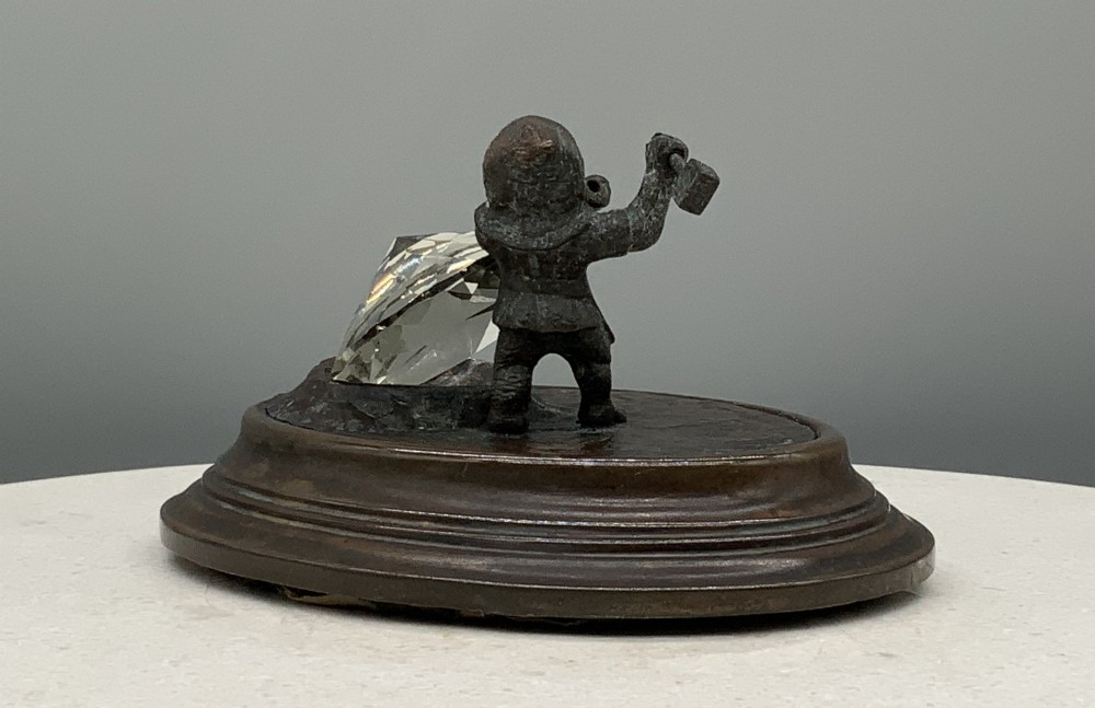 A small dark bronze figurine of a bearded dwarf miner holding a hammer overhead, standing on a molded base next to a faceted cut crystal gem. View VIII