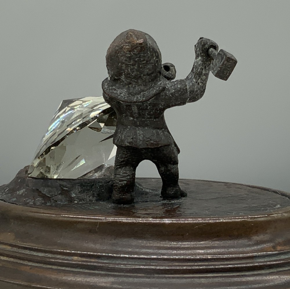 A small dark bronze figurine of a bearded dwarf miner holding a hammer overhead, standing on a molded base next to a faceted cut crystal gem. View IX