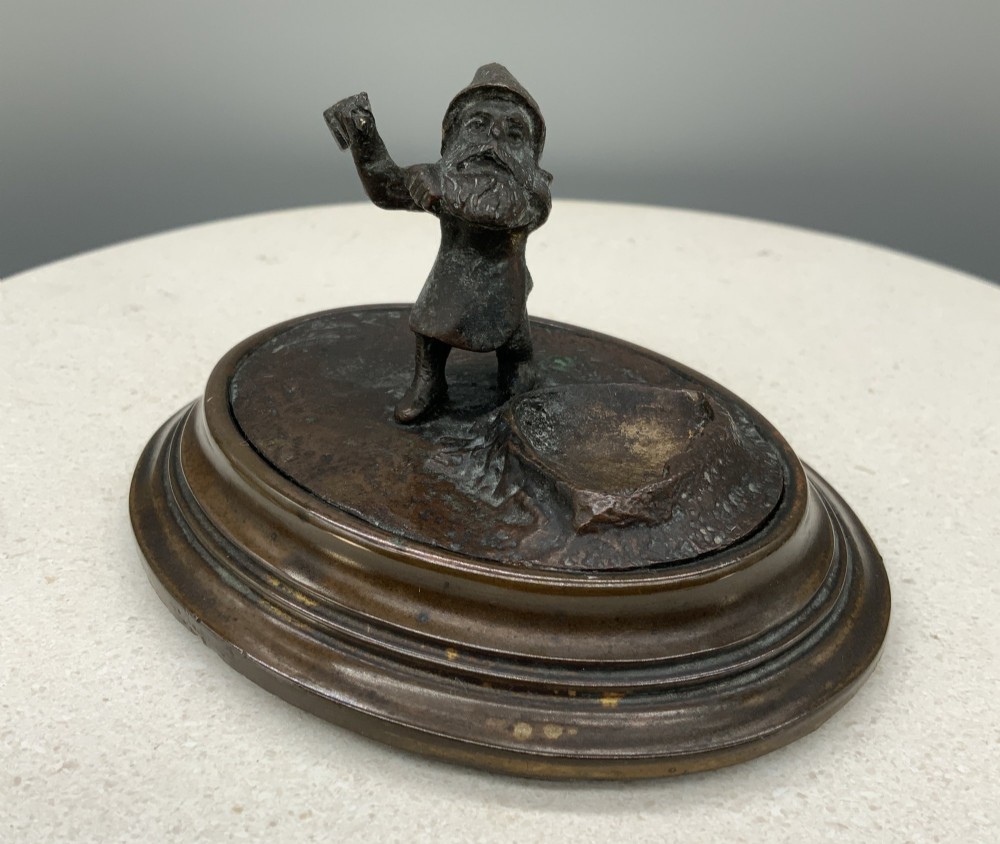 A small dark bronze figurine of a bearded dwarf miner holding a hammer overhead, standing on a molded base next to a faceted cut crystal gem. View XI