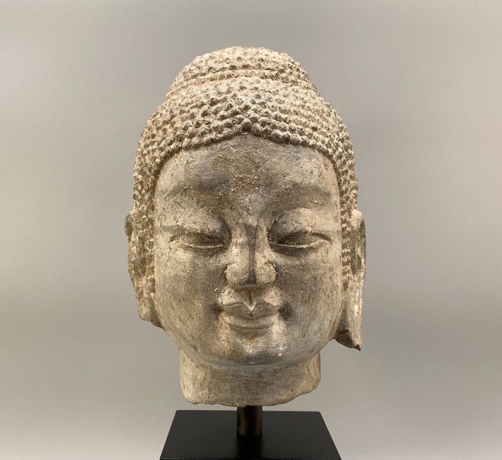 A carved limestone Chinese Buddha head featuring downcast eyes, elongated earlobes, and coiled hair, mounted on a black square stand. View IV