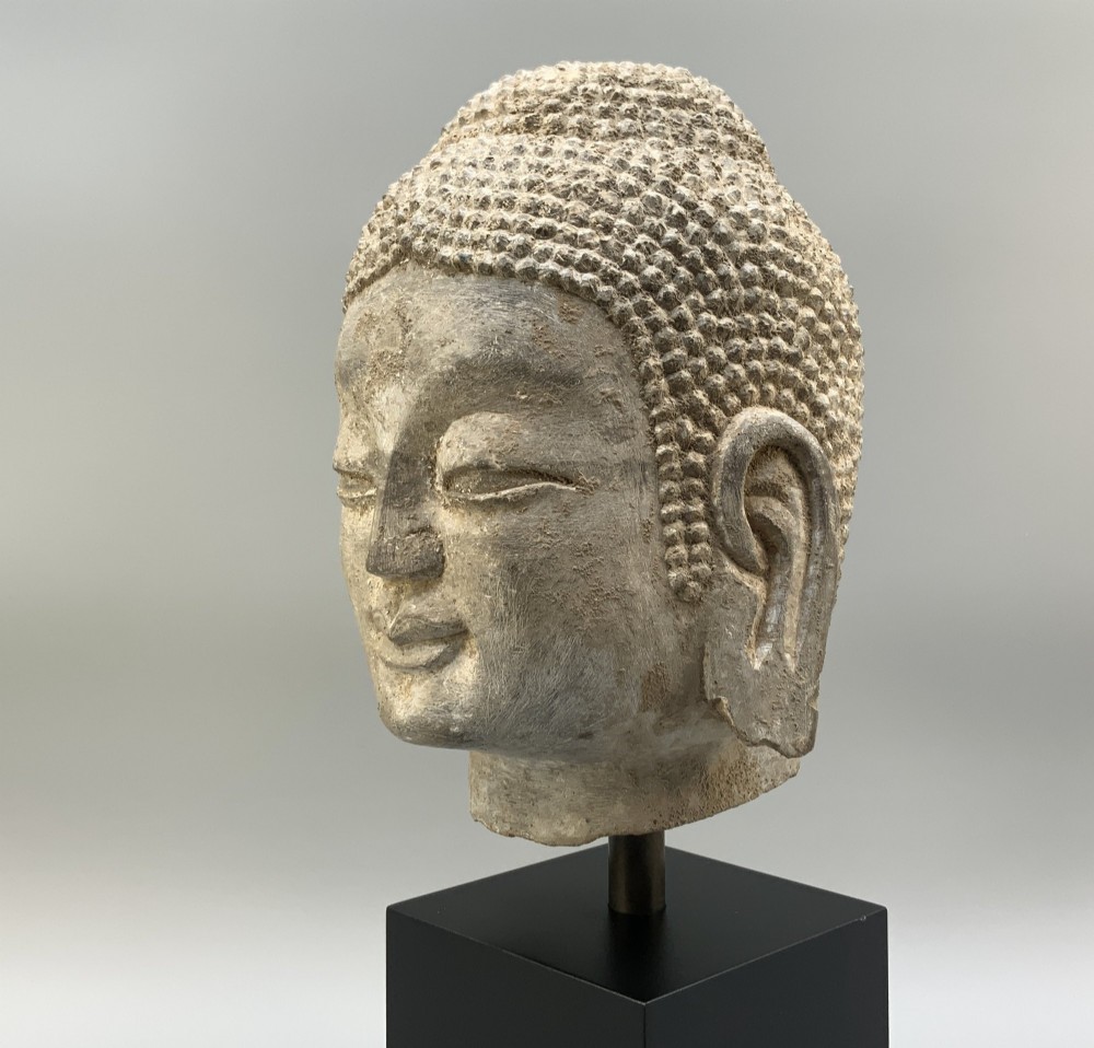 A carved limestone Chinese Buddha head featuring downcast eyes, elongated earlobes, and coiled hair, mounted on a black square stand. View VIII