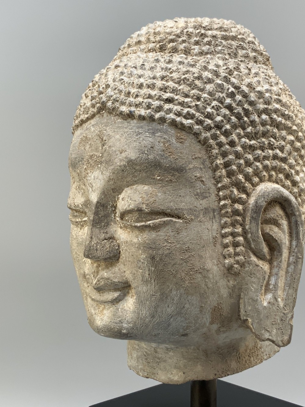 A carved limestone Chinese Buddha head featuring downcast eyes, elongated earlobes, and coiled hair, mounted on a black square stand. View IX