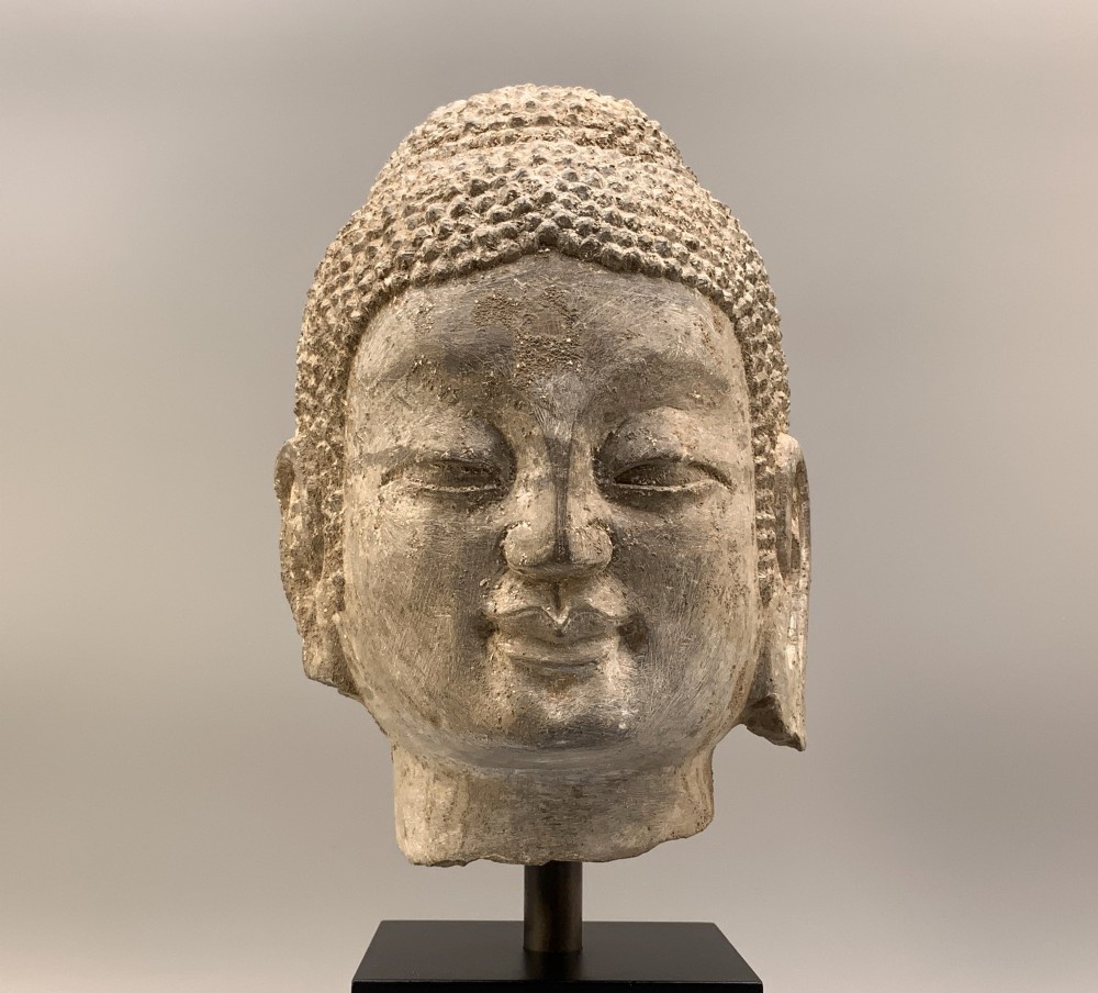 A carved limestone Chinese Buddha head featuring downcast eyes, elongated earlobes, and coiled hair, mounted on a black square stand. View X