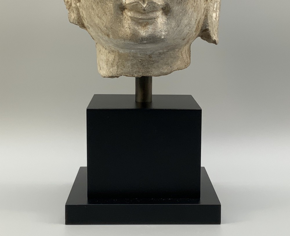 A carved limestone Chinese Buddha head featuring downcast eyes, elongated earlobes, and coiled hair, mounted on a black square stand. View XIII