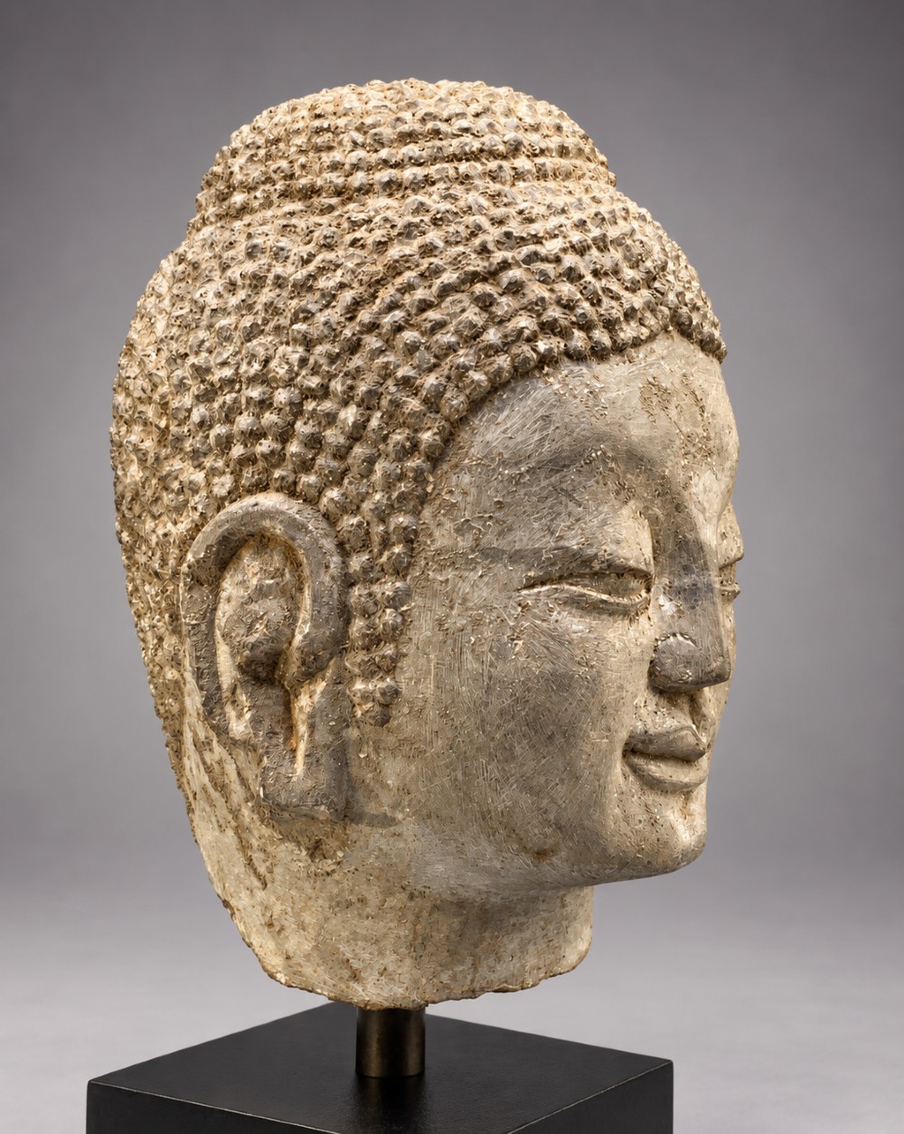 A carved limestone Chinese Buddha head featuring downcast eyes, elongated earlobes, and coiled hair, mounted on a black square stand.
