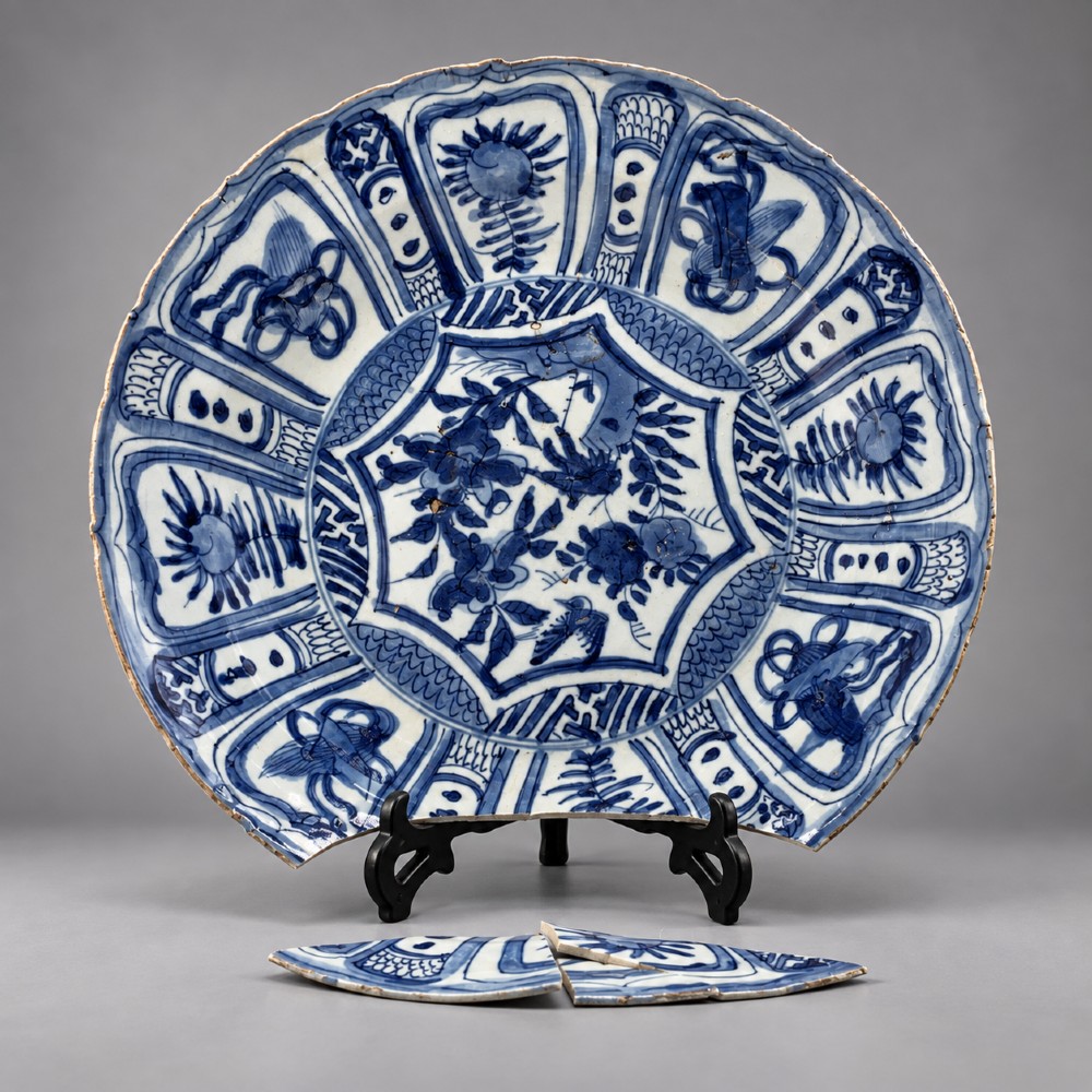 A large 17th-century Japanese Edo period blue and white porcelain charger featuring panelled floral patterns with detached rim fragments in the foreground.