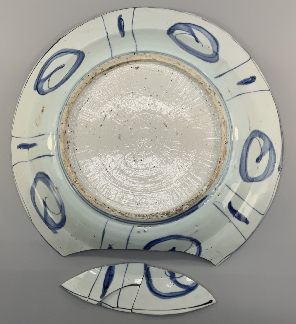 A large 17th-century Japanese Edo period blue and white porcelain charger featuring panelled floral patterns with detached rim fragments in the foreground. View X