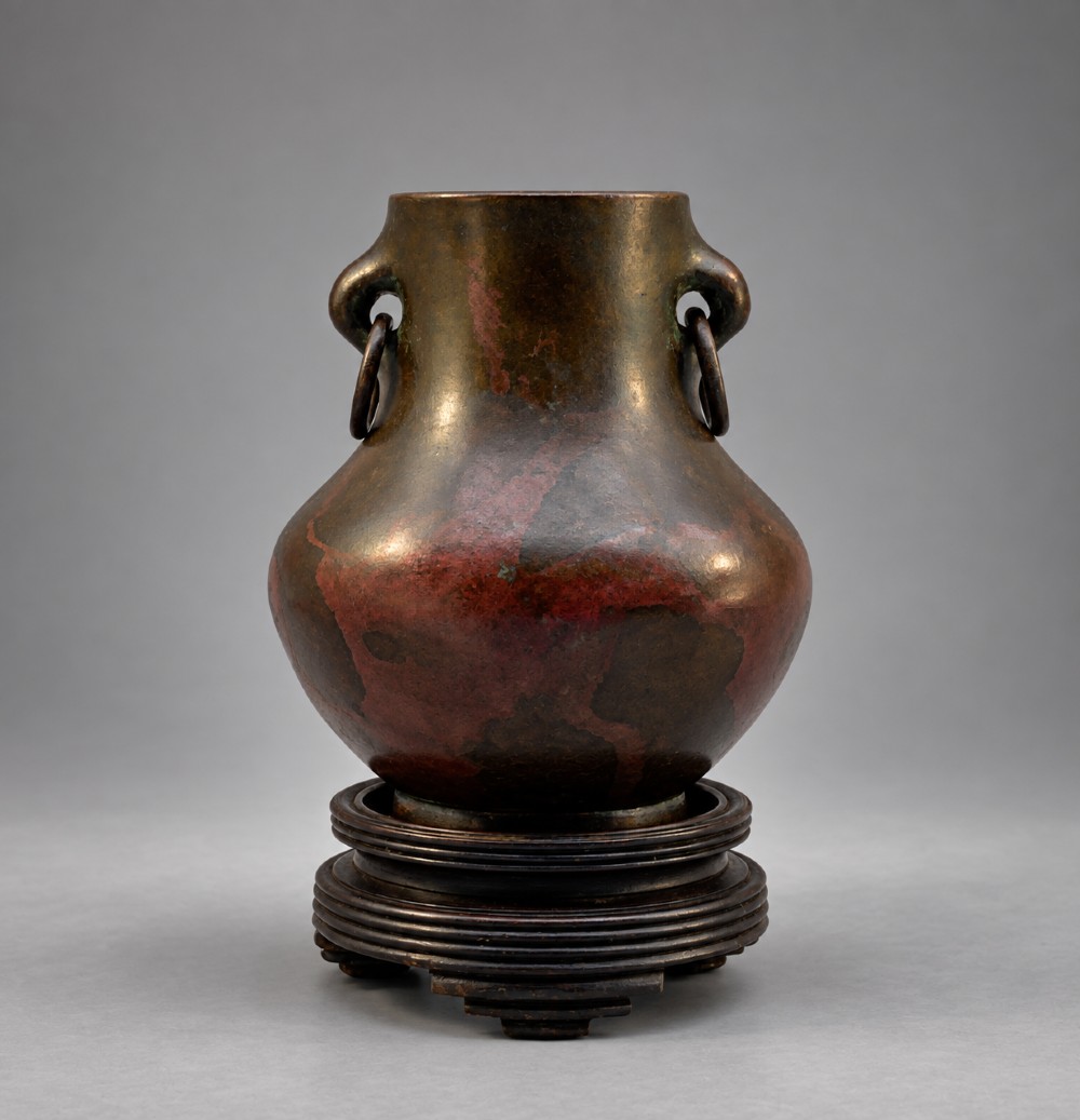 A small pear-shaped dark bronze vase with loop handles and loose rings, featuring mottled red surface patches, resting on a carved dark wooden base.