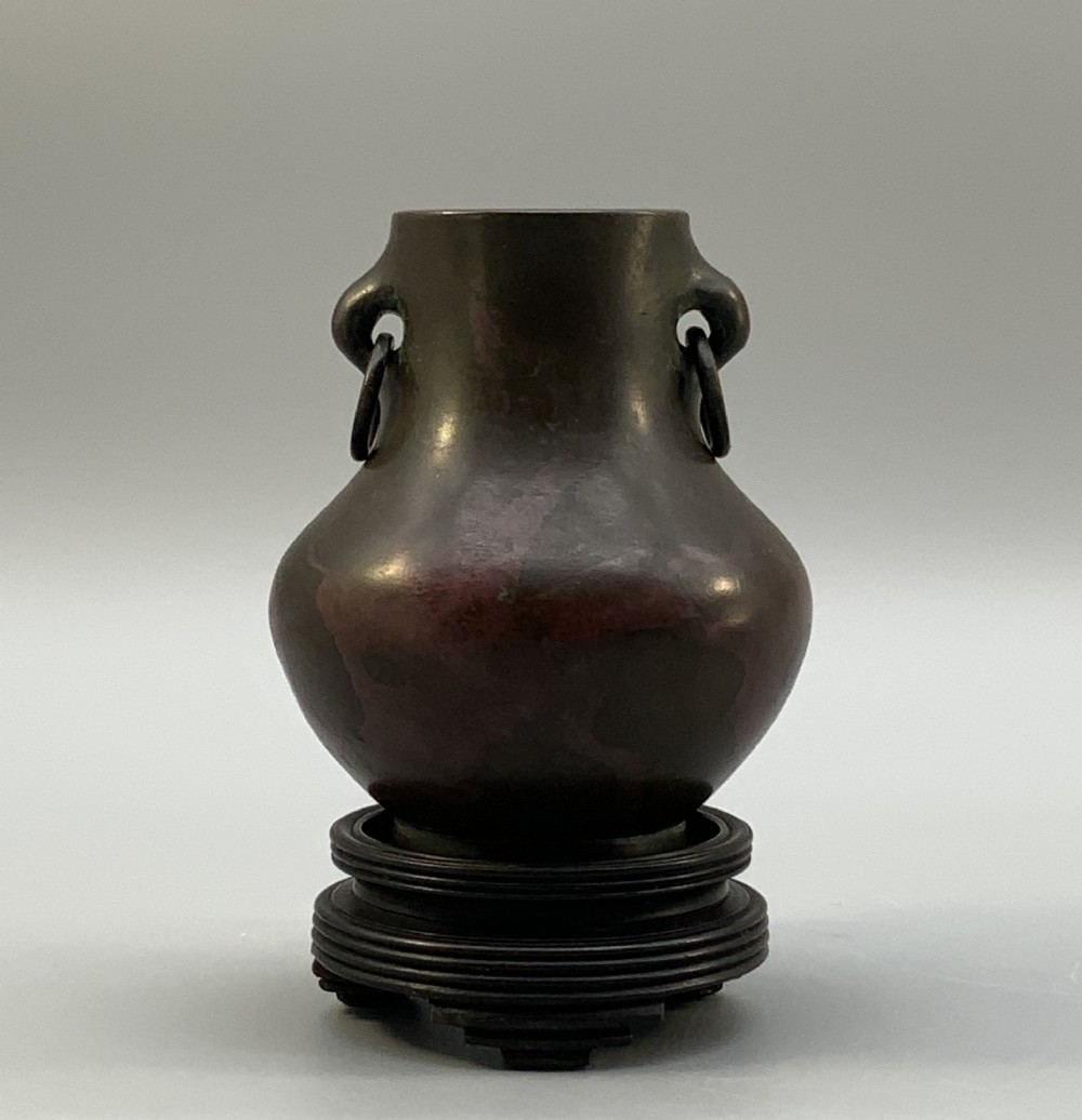 A small pear-shaped dark bronze vase with loop handles and loose rings, featuring mottled red surface patches, resting on a carved dark wooden base. View II