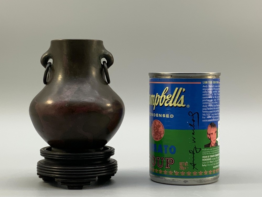 A small pear-shaped dark bronze vase with loop handles and loose rings, featuring mottled red surface patches, resting on a carved dark wooden base. View III