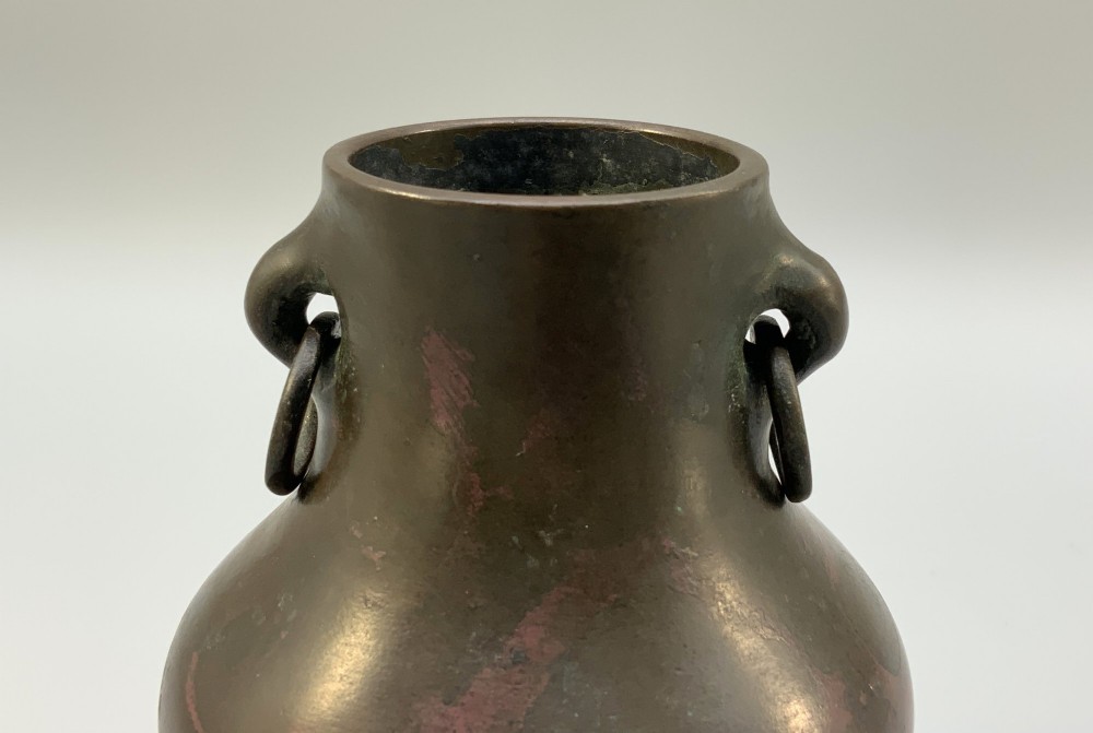 A small pear-shaped dark bronze vase with loop handles and loose rings, featuring mottled red surface patches, resting on a carved dark wooden base. View IV
