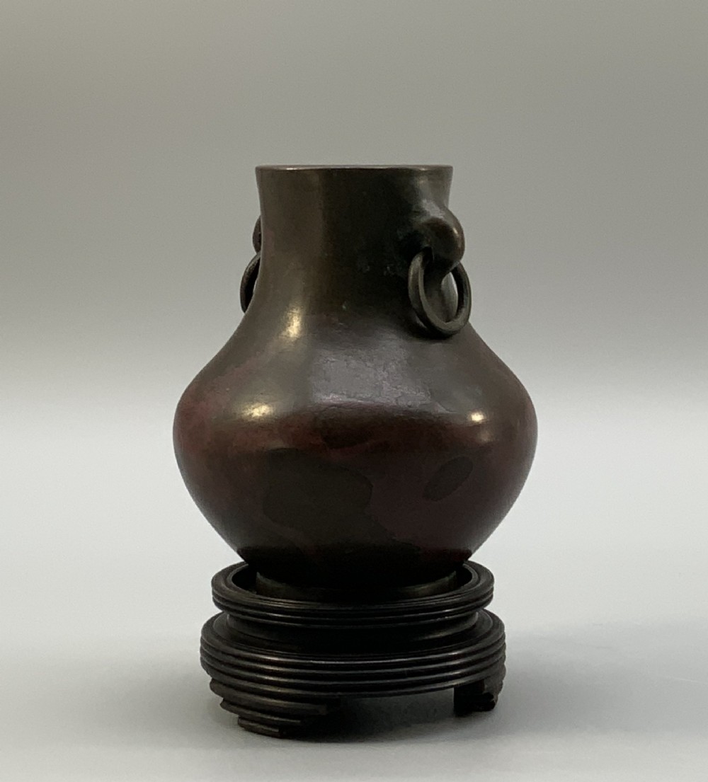 A small pear-shaped dark bronze vase with loop handles and loose rings, featuring mottled red surface patches, resting on a carved dark wooden base. View V