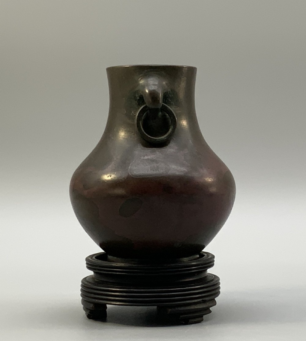 A small pear-shaped dark bronze vase with loop handles and loose rings, featuring mottled red surface patches, resting on a carved dark wooden base. View VI
