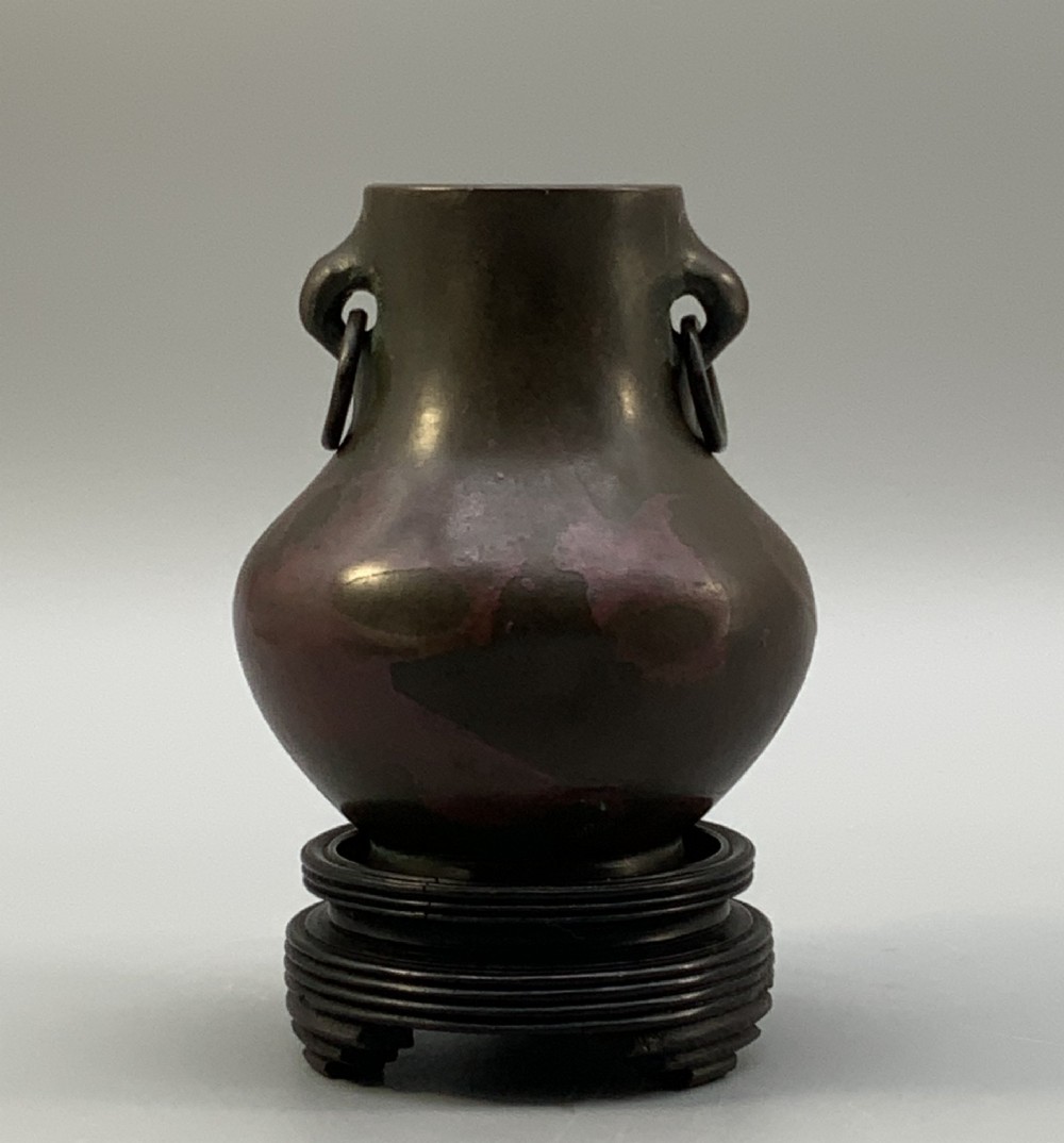 A small pear-shaped dark bronze vase with loop handles and loose rings, featuring mottled red surface patches, resting on a carved dark wooden base. View VII