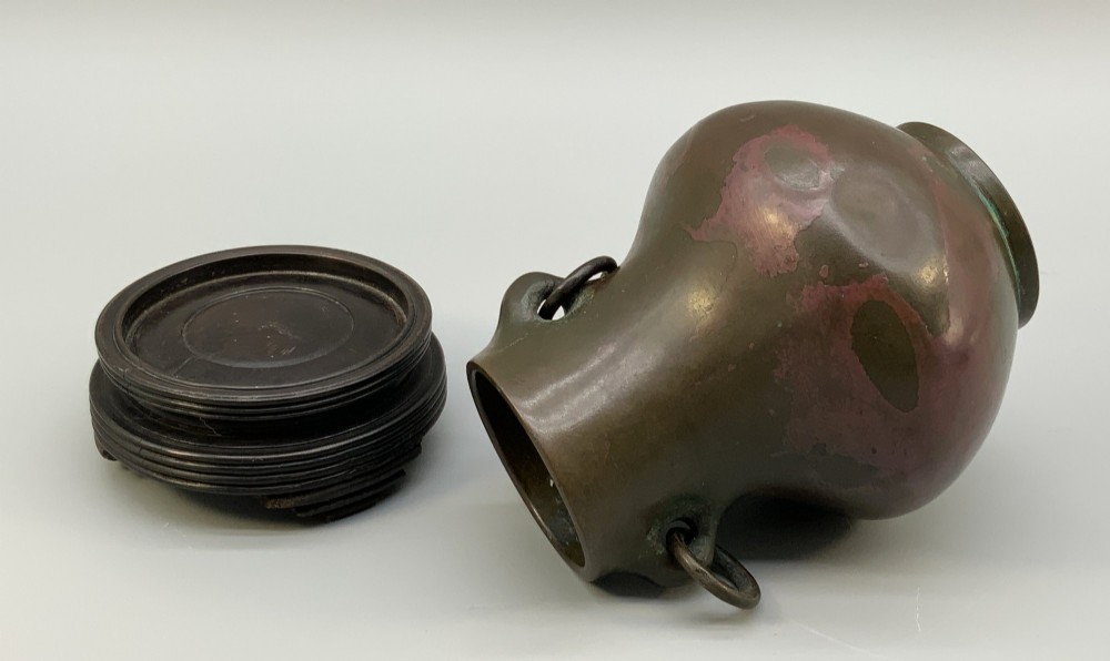 A small pear-shaped dark bronze vase with loop handles and loose rings, featuring mottled red surface patches, resting on a carved dark wooden base. View VIII