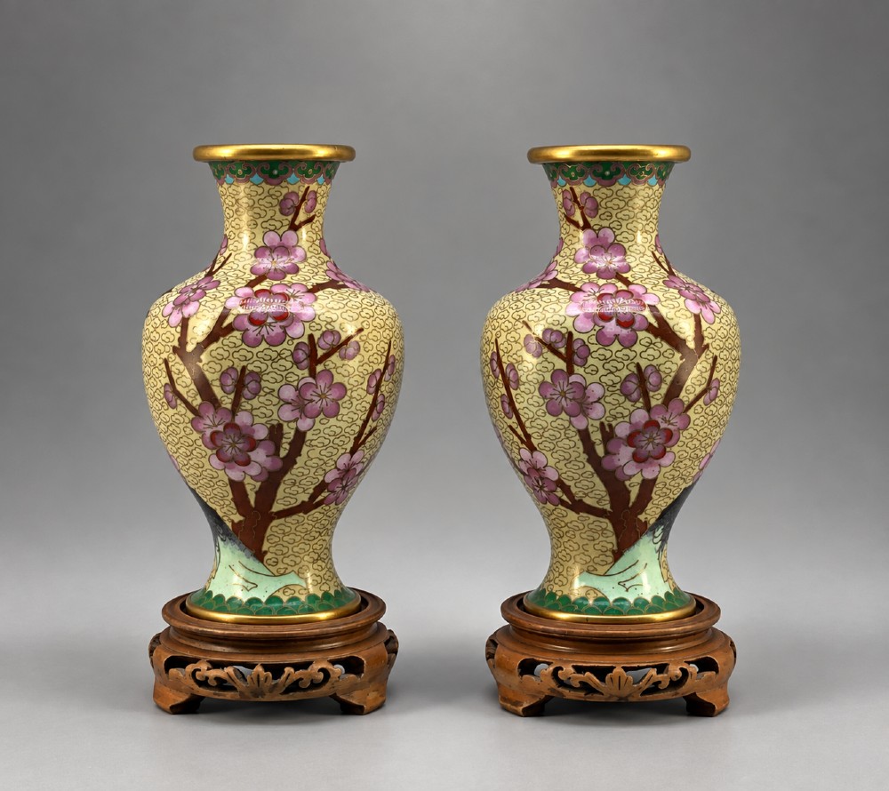 A pair of early 20th-century Chinese cloisonne enamel vases featuring pink prunus blossoms and bird motifs on a yellow cloud-scroll ground, resting on carved wooden stands.
