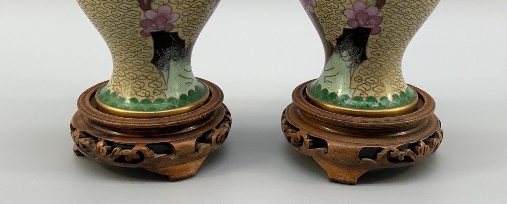 A pair of early 20th-century Chinese cloisonne enamel vases featuring pink prunus blossoms and bird motifs on a yellow cloud-scroll ground, resting on carved wooden stands. View IX