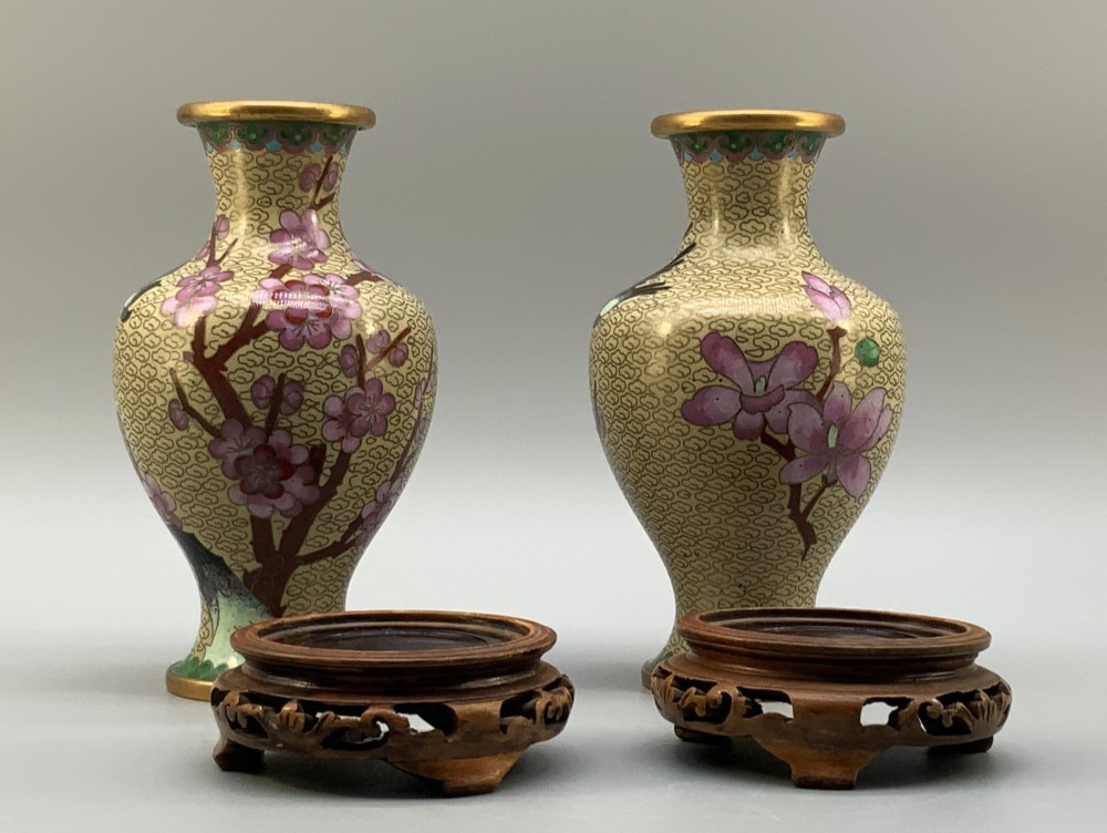 A pair of early 20th-century Chinese cloisonne enamel vases featuring pink prunus blossoms and bird motifs on a yellow cloud-scroll ground, resting on carved wooden stands. View X