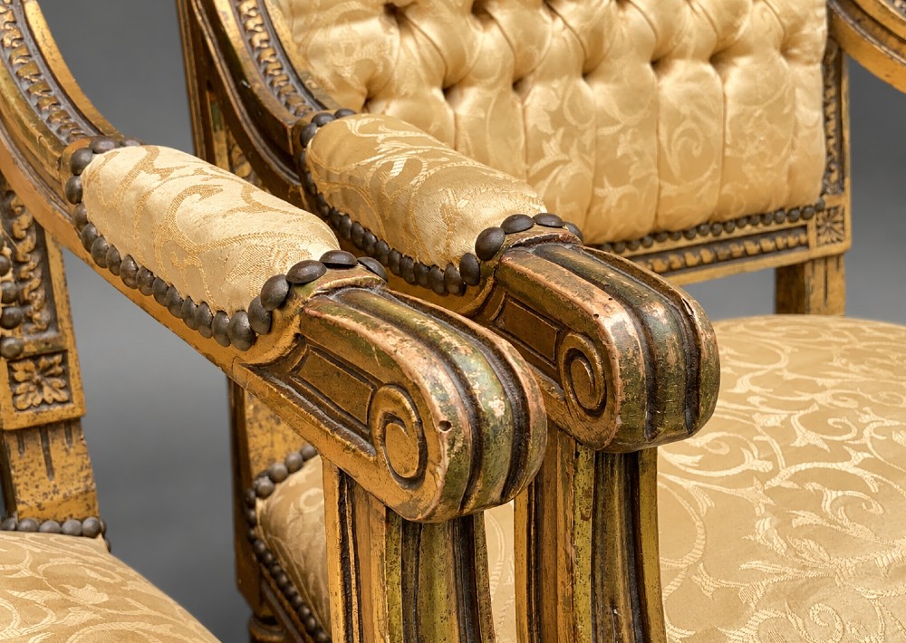 A pair of 19th-century giltwood open armchairs featuring carved leaf detailing, button-tufted gold damask upholstery, and fluted front legs. View X
