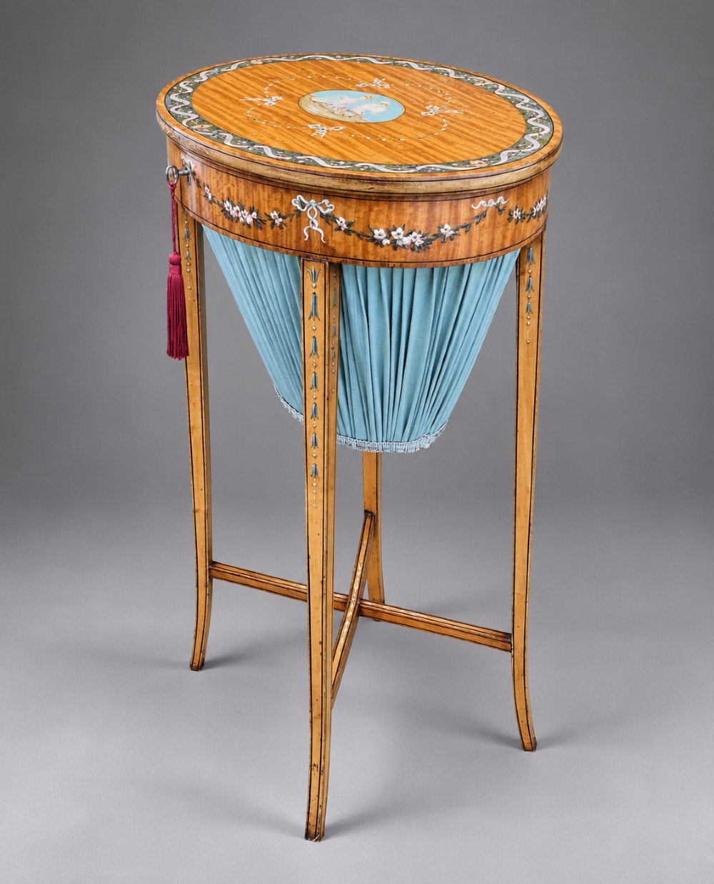 An oval satinwood sewing table featuring hand-painted floral swags, slender tapered legs, an X-form stretcher, and a pleated light blue fabric work basket suspended below.