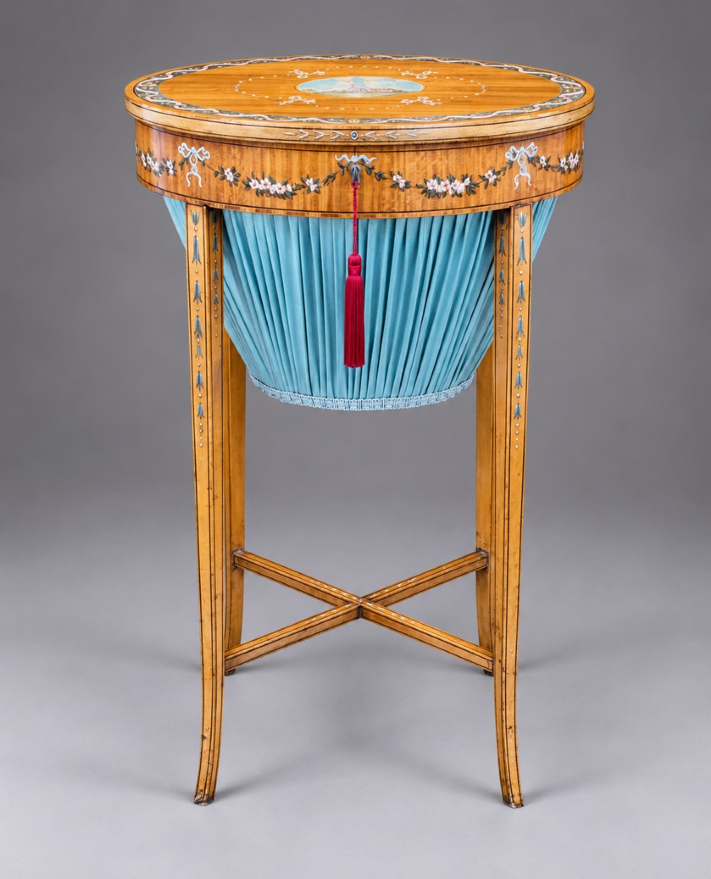 An oval satinwood sewing table featuring hand-painted floral swags, slender tapered legs, an X-form stretcher, and a pleated light blue fabric work basket suspended below. View II