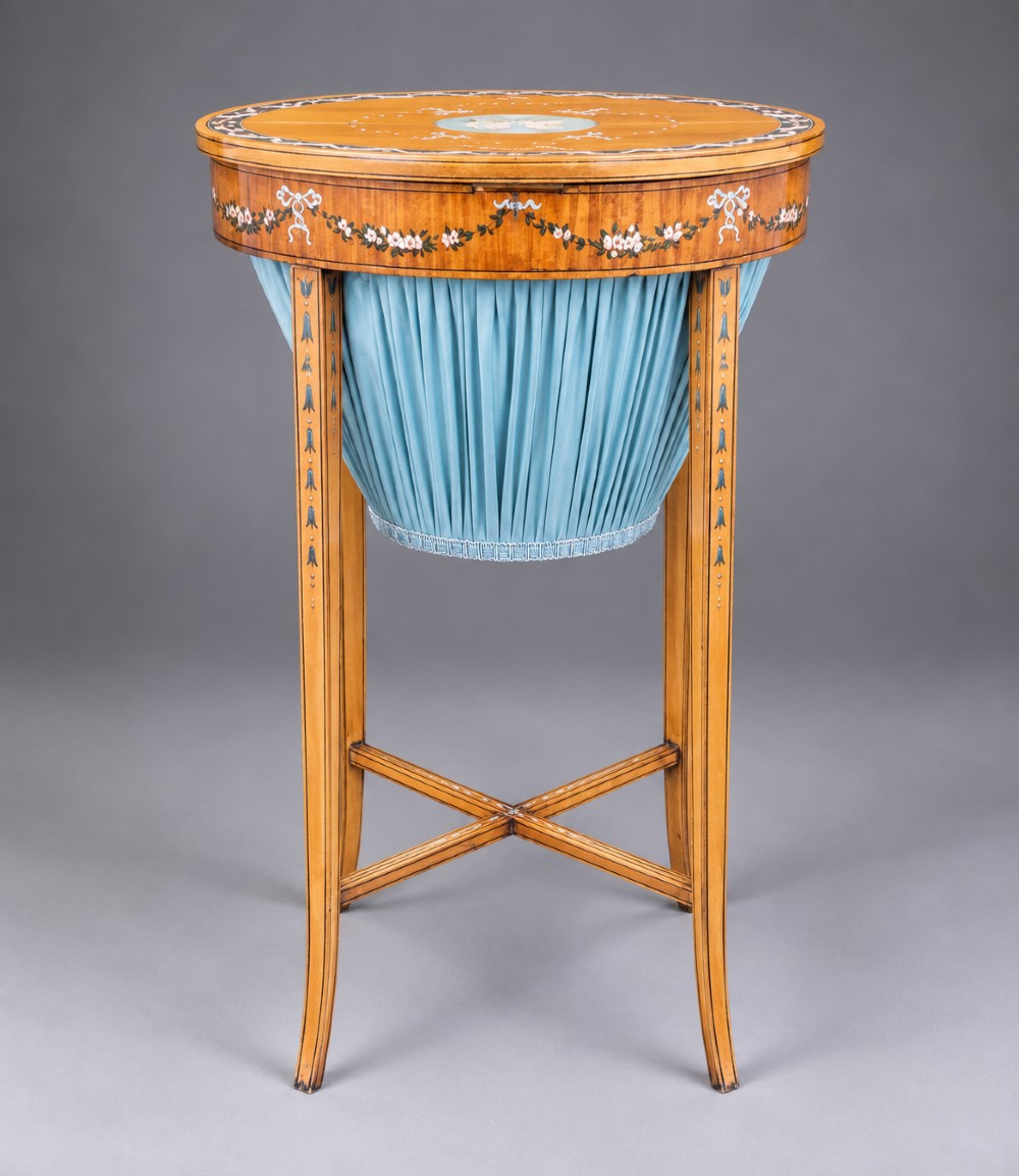 An oval satinwood sewing table featuring hand-painted floral swags, slender tapered legs, an X-form stretcher, and a pleated light blue fabric work basket suspended below. View IV