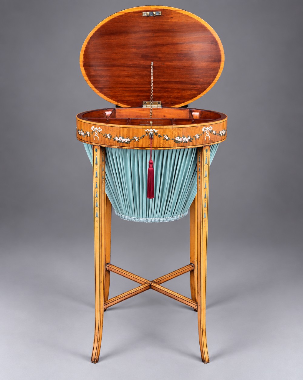 An oval satinwood sewing table featuring hand-painted floral swags, slender tapered legs, an X-form stretcher, and a pleated light blue fabric work basket suspended below. View V