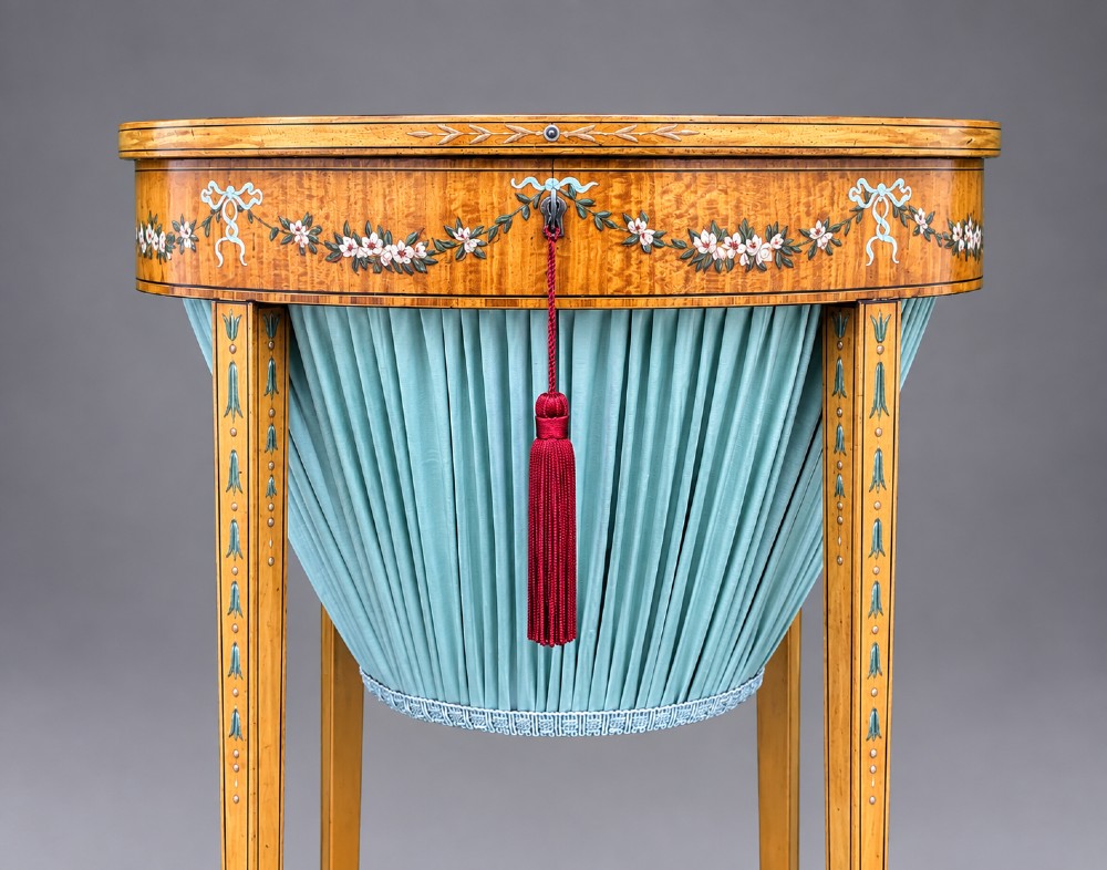 An oval satinwood sewing table featuring hand-painted floral swags, slender tapered legs, an X-form stretcher, and a pleated light blue fabric work basket suspended below. View VI