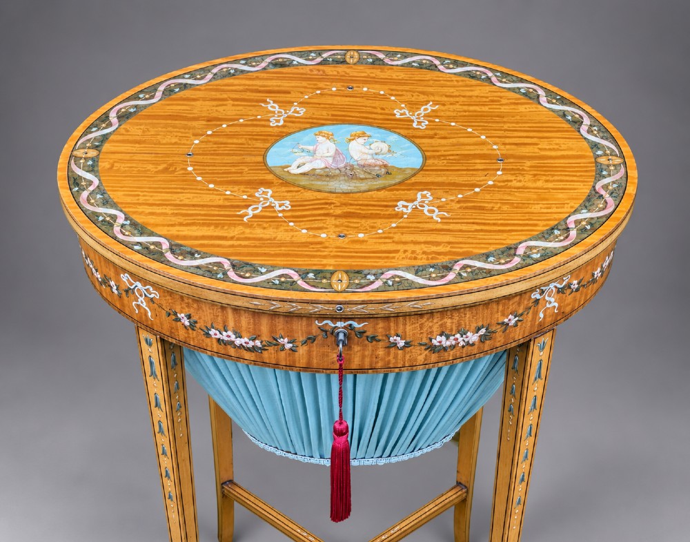 An oval satinwood sewing table featuring hand-painted floral swags, slender tapered legs, an X-form stretcher, and a pleated light blue fabric work basket suspended below. View VII