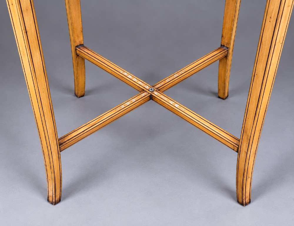 An oval satinwood sewing table featuring hand-painted floral swags, slender tapered legs, an X-form stretcher, and a pleated light blue fabric work basket suspended below. View X