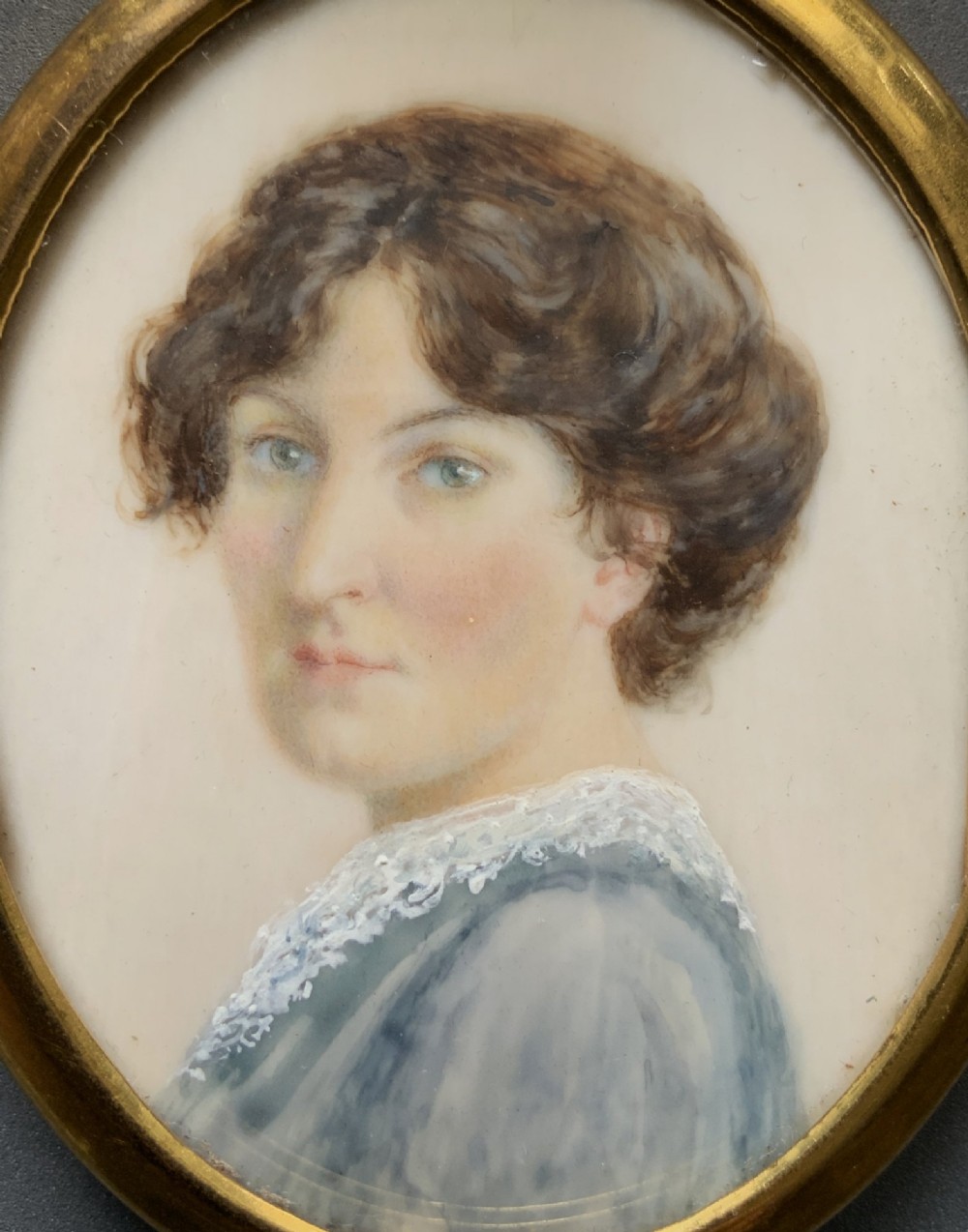 An Edwardian oval watercolour portrait miniature depicting a lady with dark curly hair and a lace-collared blue dress, set within a gilt brass frame with a hanging ring. View II