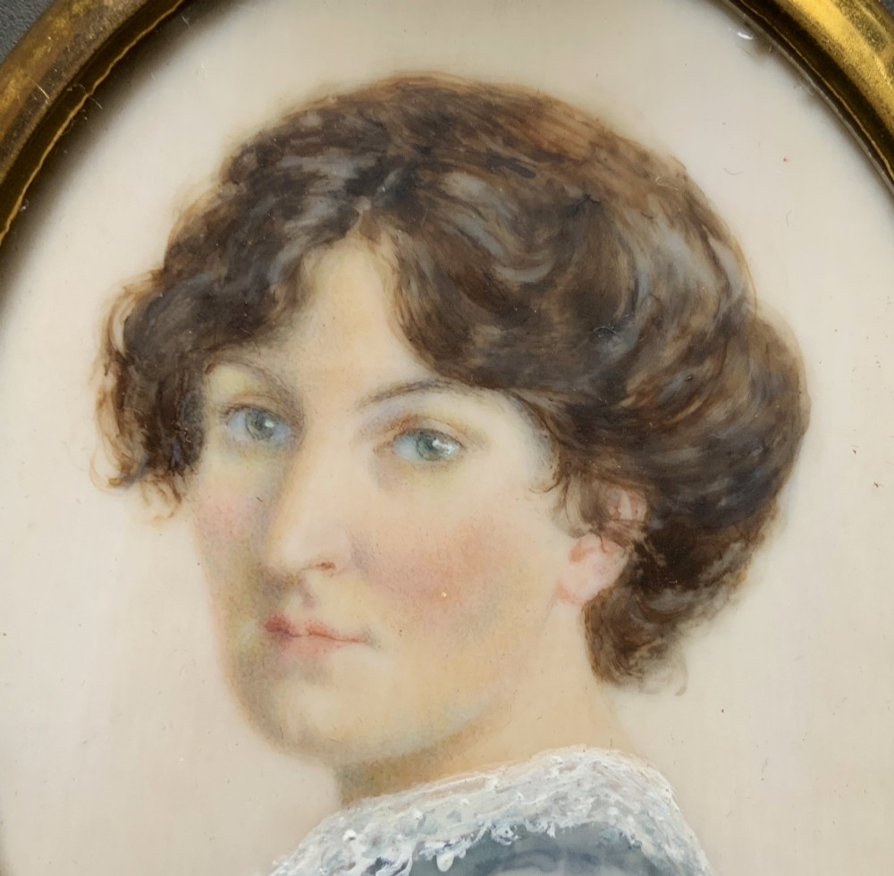 An Edwardian oval watercolour portrait miniature depicting a lady with dark curly hair and a lace-collared blue dress, set within a gilt brass frame with a hanging ring. View IV