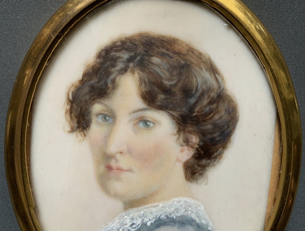 An Edwardian oval watercolour portrait miniature depicting a lady with dark curly hair and a lace-collared blue dress, set within a gilt brass frame with a hanging ring. View V