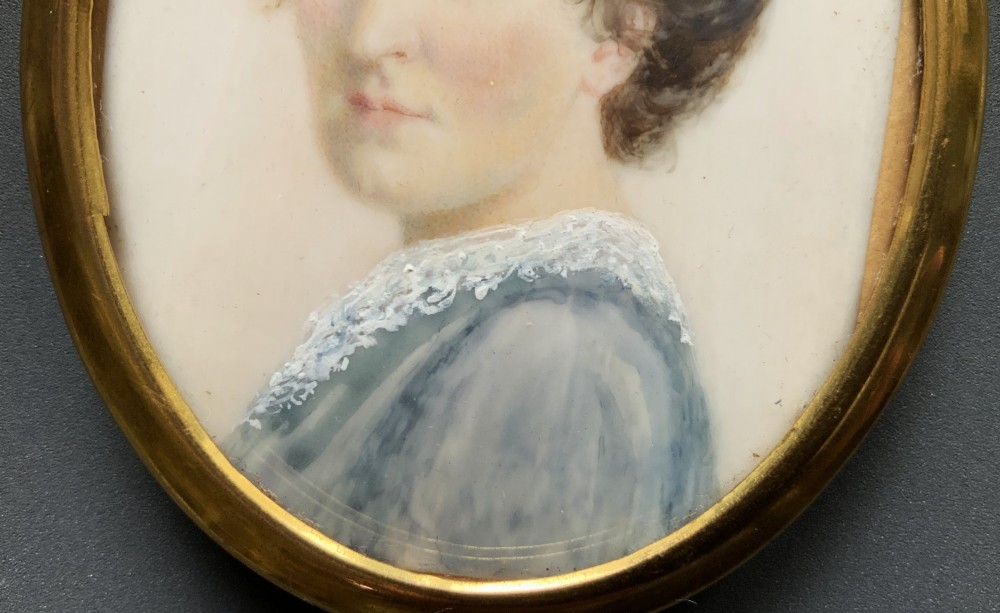 An Edwardian oval watercolour portrait miniature depicting a lady with dark curly hair and a lace-collared blue dress, set within a gilt brass frame with a hanging ring. View VI