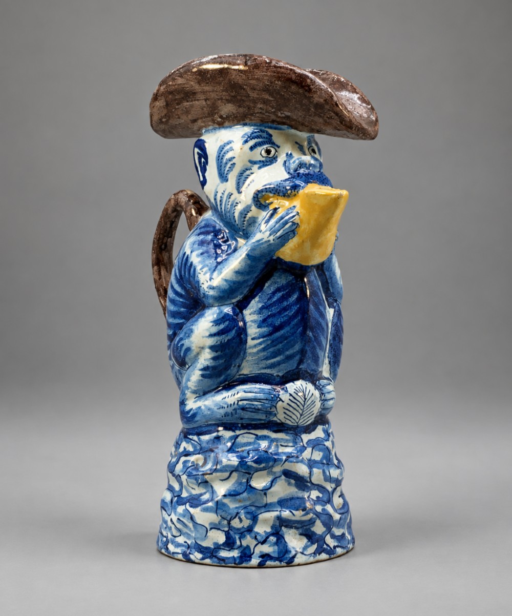 An 18th-century Dutch Delftware ceramic jug hand-painted in blue, white, brown, and yellow, designed in the form of a seated monkey in a tricorn hat holding a cup.