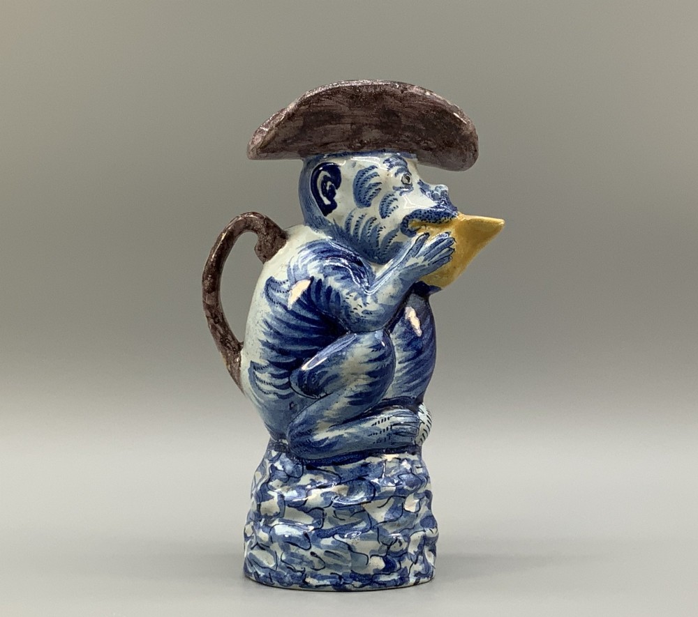 An 18th-century Dutch Delftware ceramic jug hand-painted in blue, white, brown, and yellow, designed in the form of a seated monkey in a tricorn hat holding a cup. View II