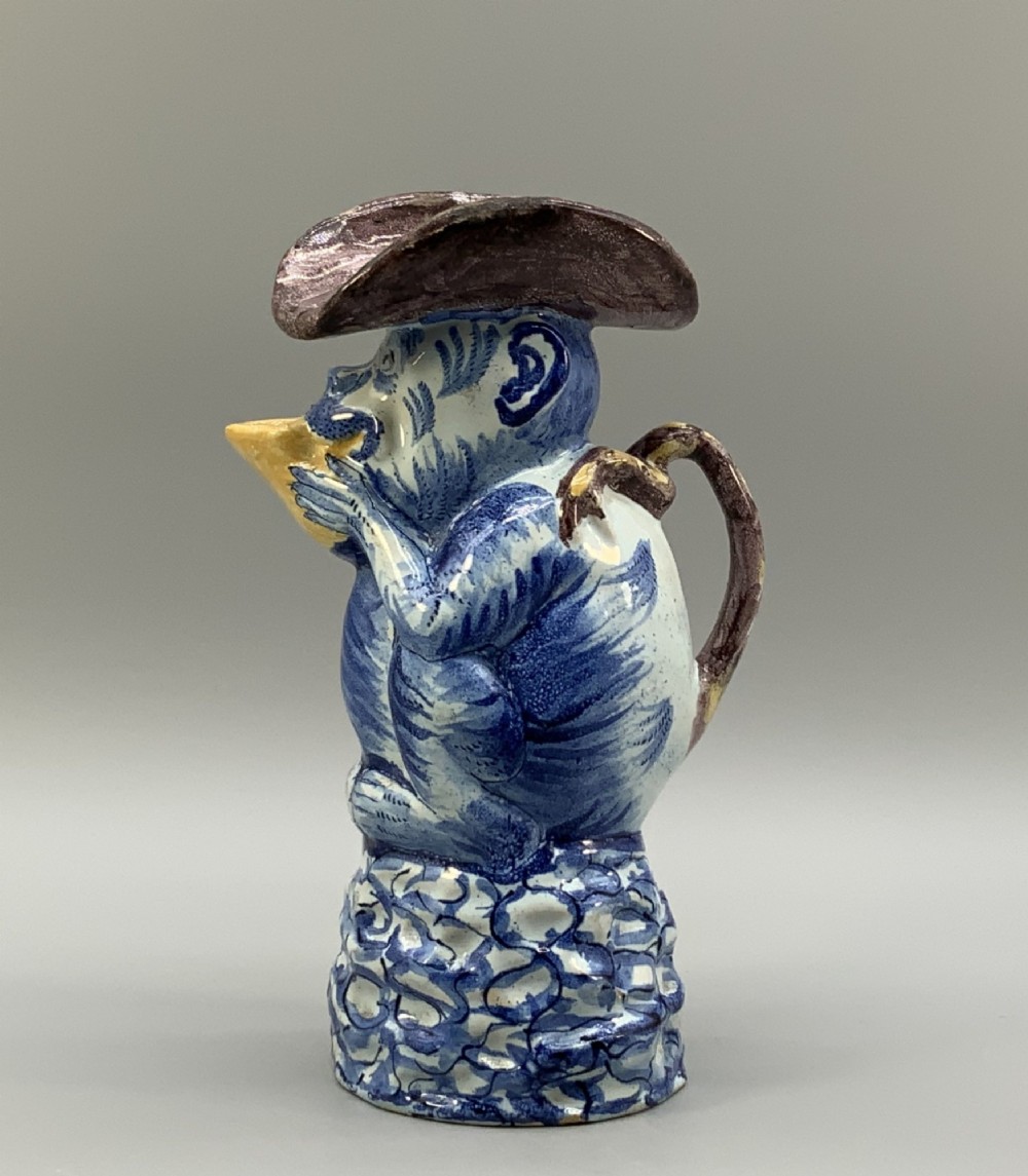 An 18th-century Dutch Delftware ceramic jug hand-painted in blue, white, brown, and yellow, designed in the form of a seated monkey in a tricorn hat holding a cup. View IV