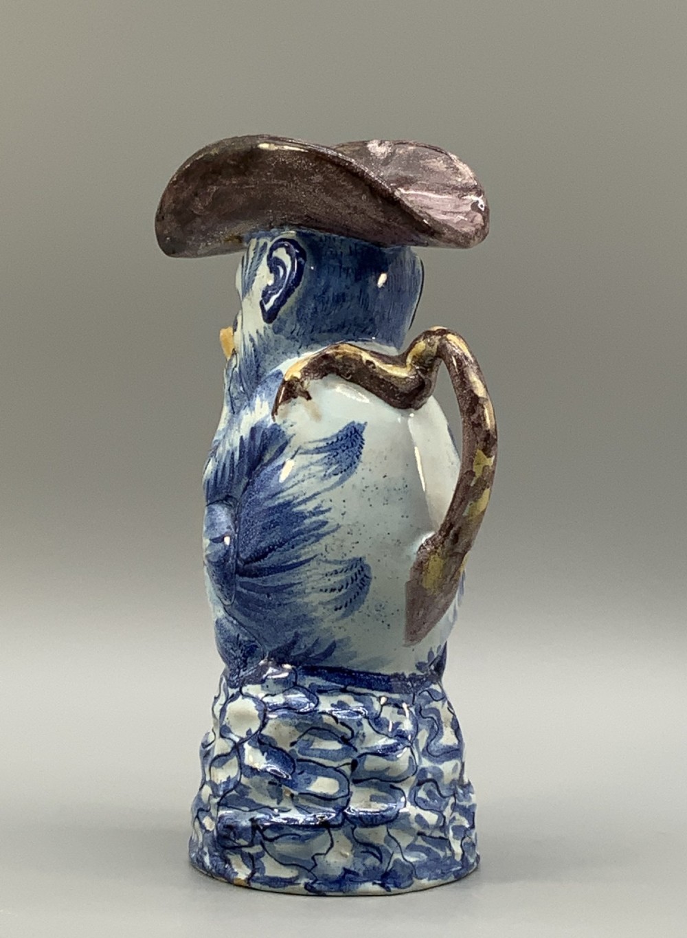 An 18th-century Dutch Delftware ceramic jug hand-painted in blue, white, brown, and yellow, designed in the form of a seated monkey in a tricorn hat holding a cup. View V