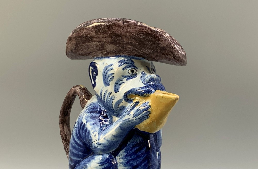 An 18th-century Dutch Delftware ceramic jug hand-painted in blue, white, brown, and yellow, designed in the form of a seated monkey in a tricorn hat holding a cup. View VI