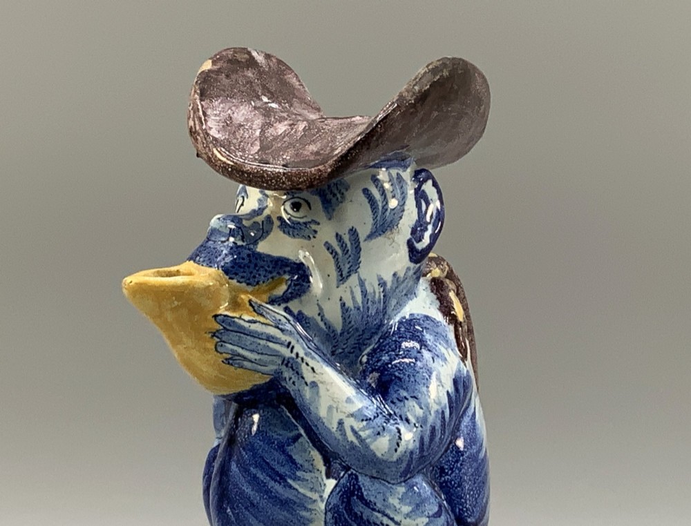 An 18th-century Dutch Delftware ceramic jug hand-painted in blue, white, brown, and yellow, designed in the form of a seated monkey in a tricorn hat holding a cup. View VIII