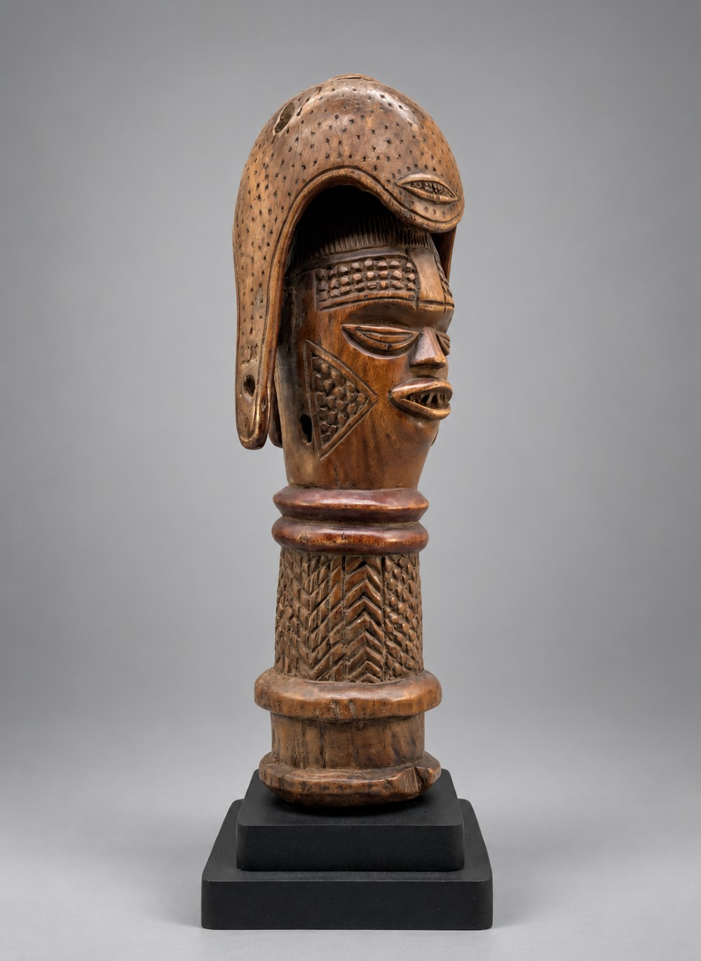 A carved brown wooden African Mende Sowei staff finial depicting a stylised head with crest, mounted on a square black plinth.