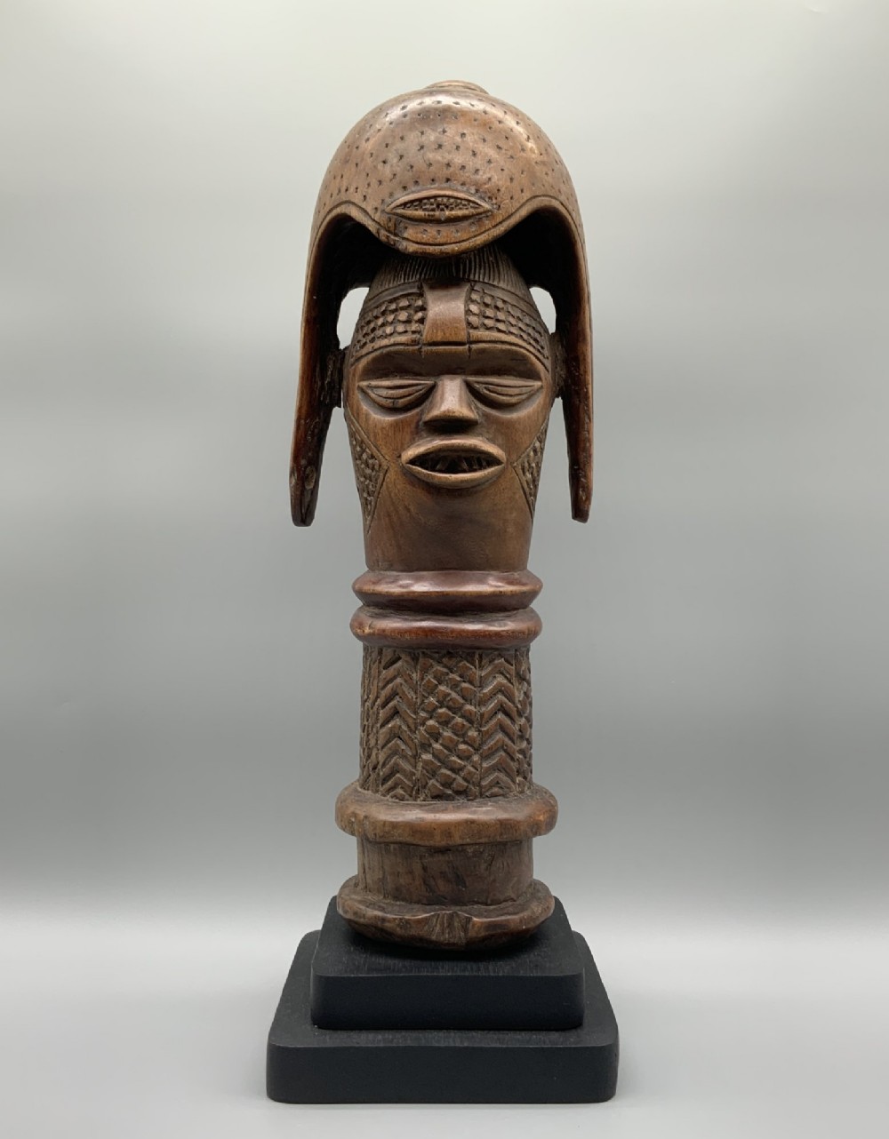 A carved brown wooden African Mende Sowei staff finial depicting a stylised head with crest, mounted on a square black plinth. View II