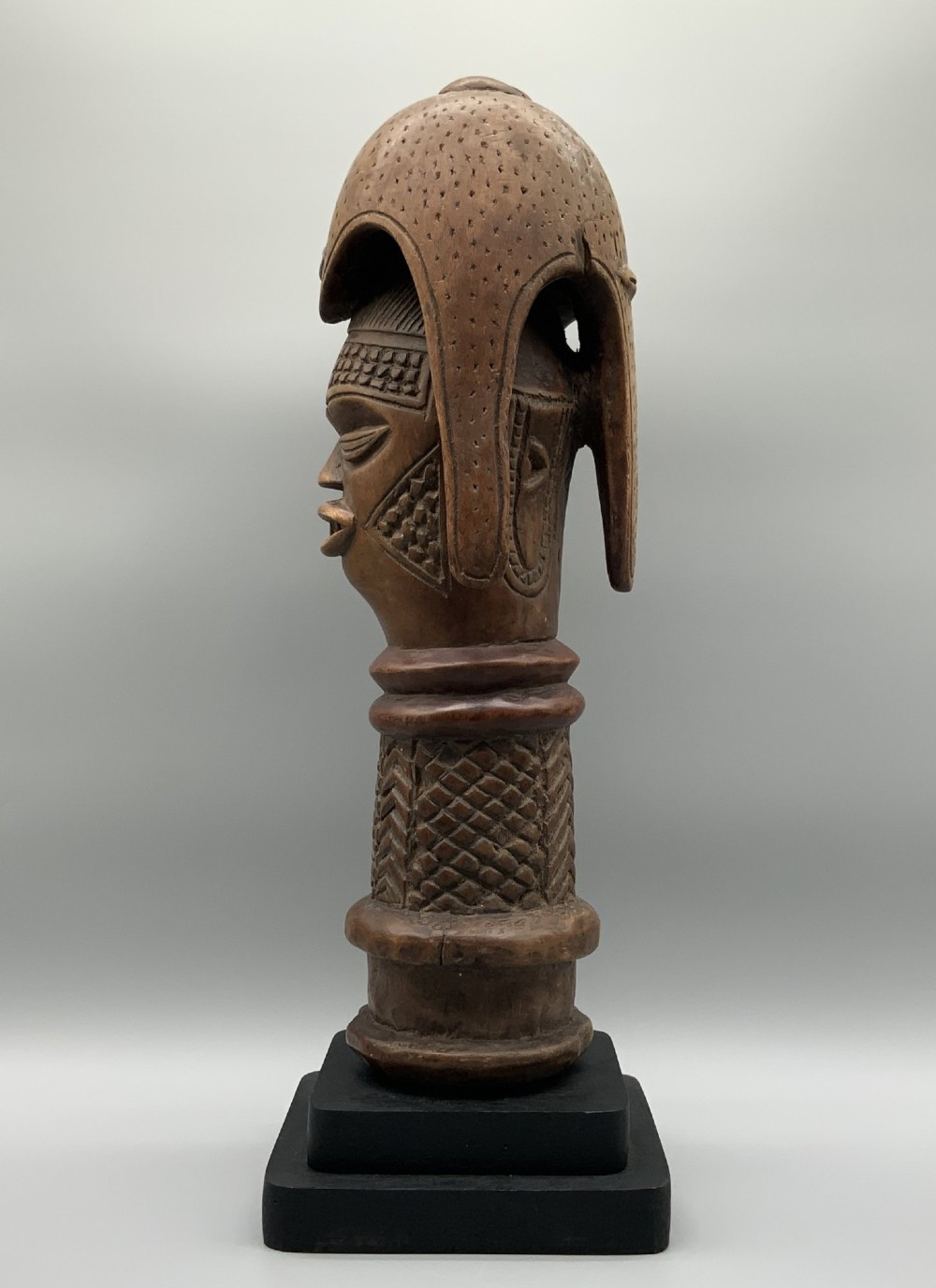 A carved brown wooden African Mende Sowei staff finial depicting a stylised head with crest, mounted on a square black plinth. View V