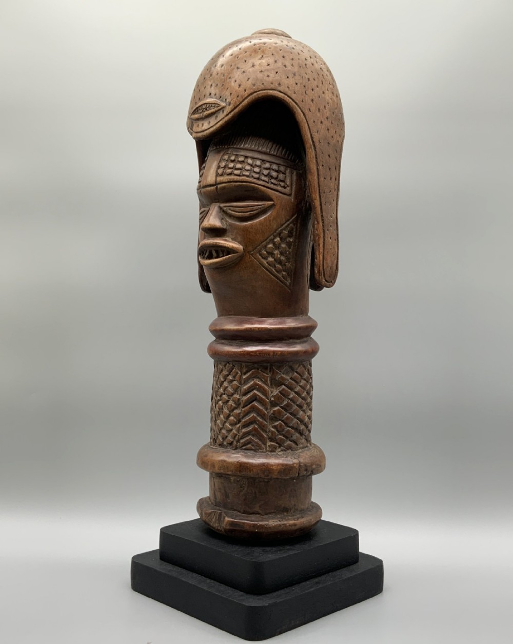 A carved brown wooden African Mende Sowei staff finial depicting a stylised head with crest, mounted on a square black plinth. View VII