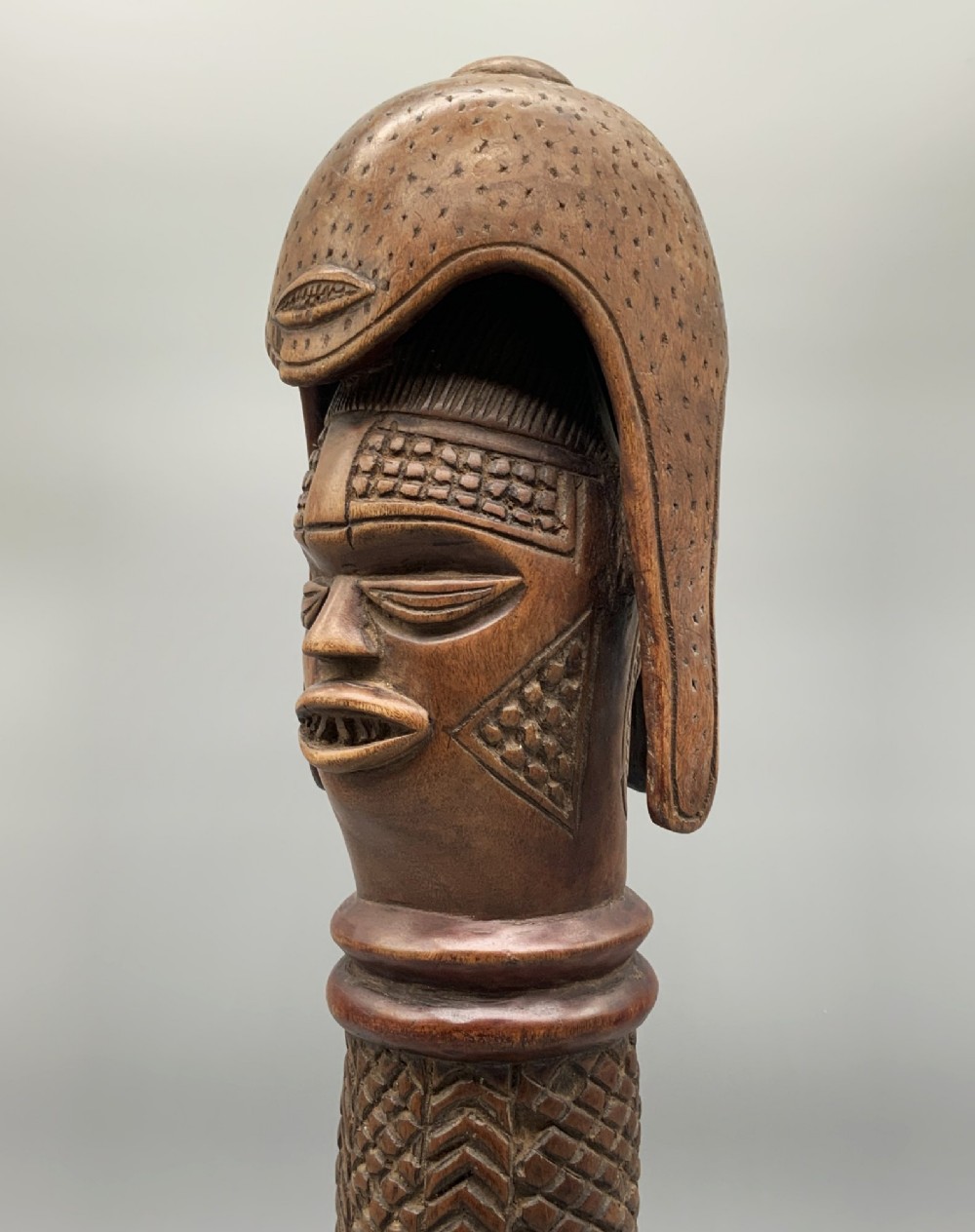 A carved brown wooden African Mende Sowei staff finial depicting a stylised head with crest, mounted on a square black plinth. View VIII