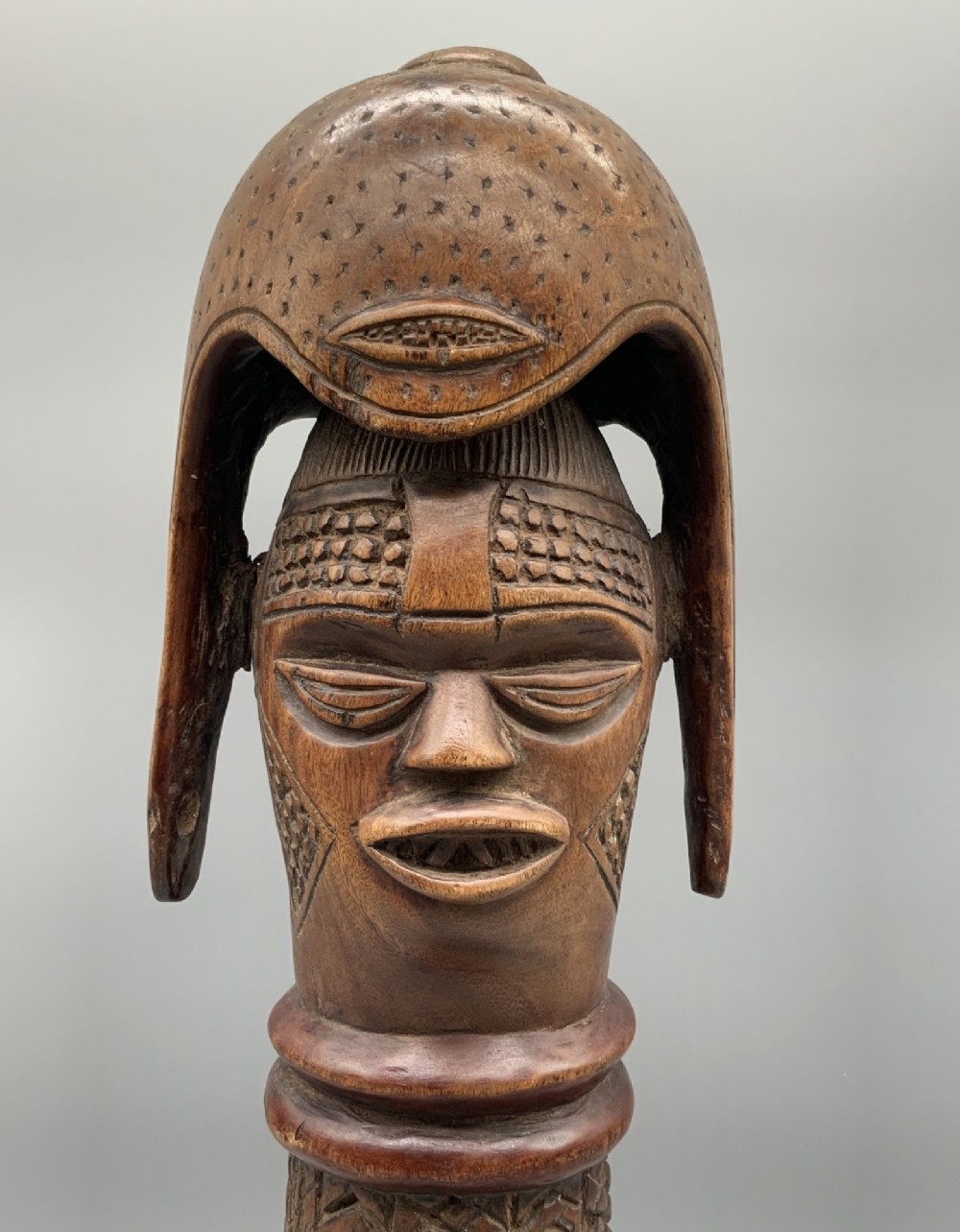 A carved brown wooden African Mende Sowei staff finial depicting a stylised head with crest, mounted on a square black plinth. View IX